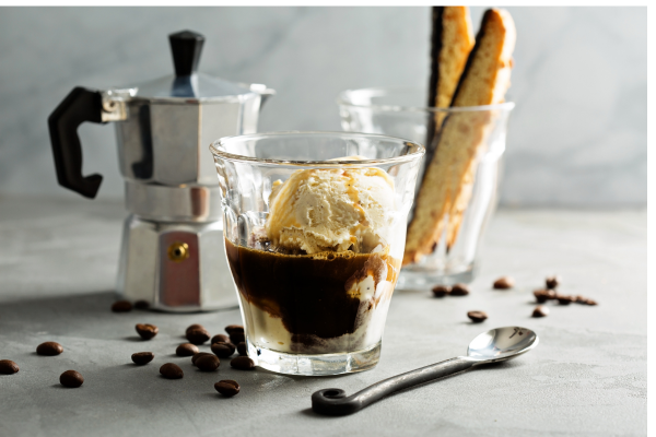 An affogato beside a moka espresso pot and glass tumbler filled with biscotti. Coffee beans are scattered around